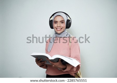 Smiling Asian Muslim female student in pink sweater with bag while listening music, using headset and reading book isolated on white background. back to school concept