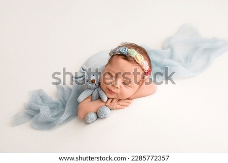 A newborn baby girl in a flower headband sleeps with a blue rabbit in her hands. The girl lies on white background covered with a light blue blanket. Royalty-Free Stock Photo #2285772357