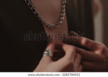 Female hands, silver chain hanging around the neck of a woman dressed in a black jacket. Cropped figure, no face.
