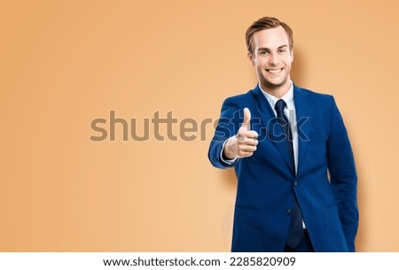 Image of excited businessman showing thumbs up like hand sign gesture, in blue suit, over latte beige background. Handsome happy man. Copy space for ad.