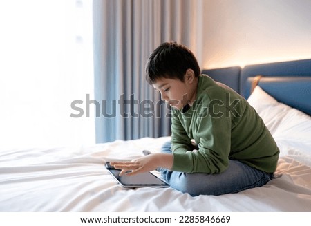 Kid boy using tablet playing game on internet with friend, Homeschooling Kid doing homework online by digital pad at home,Child sitting on bed relaxing,watching cartoon or talking online with friend