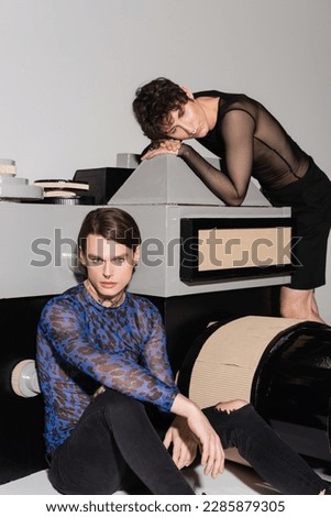 trendy nonbinary person in animal print top sitting near partner and model of photo camera on grey background