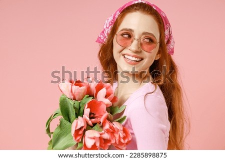 Close up young smiling happy trendy attractive woman wear rose clothes bandana glasses hold tulips flowers bouquet look aside isolated on pastel pink background studio portrait. Spring season concept