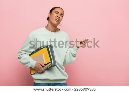 displeased african american student in sweatshirt holding gadgets and study supplies while whining isolated on pink Royalty-Free Stock Photo #2285909385