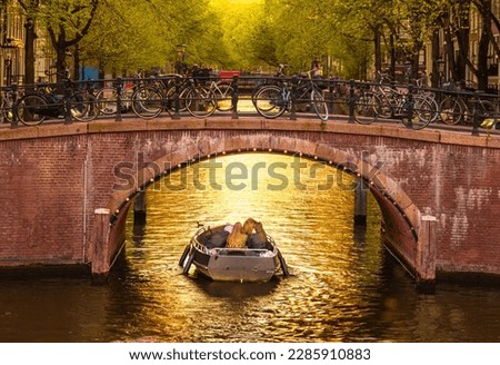 A group of young people enjoying in the boat the golden hour reflections on a canal in Amsterdam, Netherlands surrounded by stunning arch bridges and architecture.