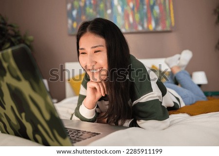 Happy asian kazachstan middle aged young girl woman laying in bed smiling wearing casual new stylish outfit smiling while watching movies cartoons series online on website. Royalty-Free Stock Photo #2285911199