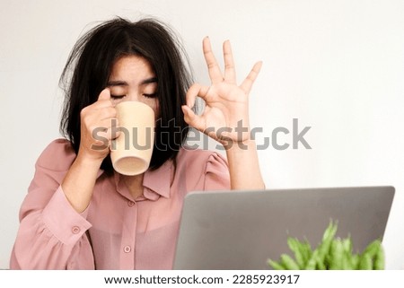 Tired Asian worker lack of sleep and energy drinking a cup of coffee and trying to work sitting at the office with a laptop on desk  Royalty-Free Stock Photo #2285923917