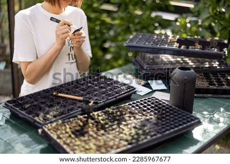 Young gardener signs the names of the plants that she plants into seedling trays in garden. Concept of hobby or small business of growing flowers