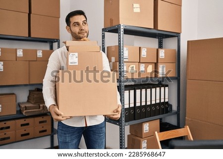 Young hispanic man with beard working at small business ecommerce holding delivery boxes smiling looking to the side and staring away thinking. 