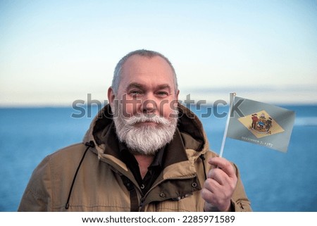 Man holding Delaware state flag. 
Portrait of older man with Delaware flag. 
Visit Delaware. Older man 50 55 60 years old 
with gray beard outdoors travelling.