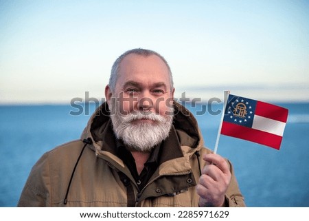 Man holding Georgia state flag. 
Portrait of older man with Georgia flag. 
Visit Georgia. Older man 50 55 60 years old 
with gray beard outdoors travelling.