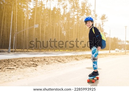 happy child schoolboy with a backpack goes to school on a skateboard. a joyful boy with protection on his knees and elbows, with a helmet on his head, stands on a plastic bright city cruiser skate