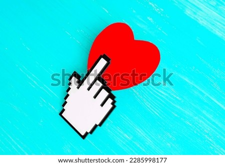 White blocky plastic pointer cursor clicks a red heart shape placed on a blue wooden background. Web and graphic design, advertising and social media related concept.
