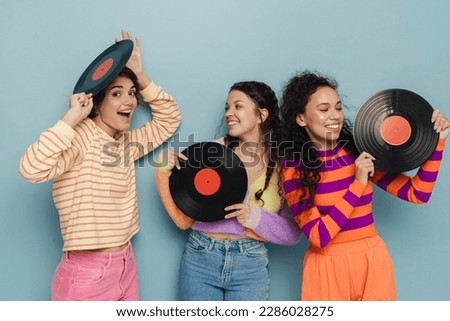 Three beautiful young girls in colorful clothes posing with vinyl records and smiling isolated over blue studio background