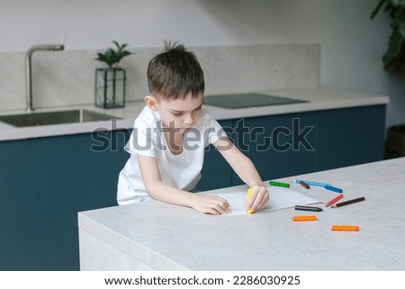 Six years old boy drawing with pencils. Modern kitchen interior in blue color. 