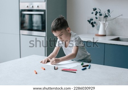 Six years old boy drawing with pencils. Modern kitchen interior in blue color. 