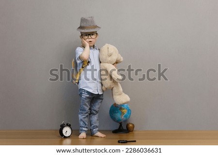 A boy in blue jeans, a blue shirt with a big toy bear and a rucksack on his back stands on the floor and is surprised at something. Little schoolboy on a gray background. High quality photo