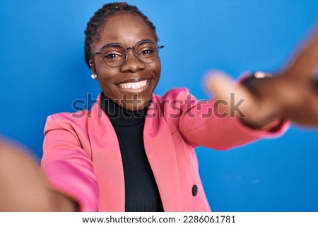 Beautiful black woman standing over blue background looking at the camera smiling with open arms for hug. cheerful expression embracing happiness. 