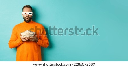 Handsome man in orange sweater and 3d glasses watching movies thrilled, holding popcorn, looking amazed, standing over blue background.