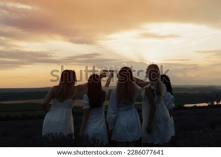 Golden evening sunbeams shine while female friends having picnic and clinking glasses with wine in lavender field. 