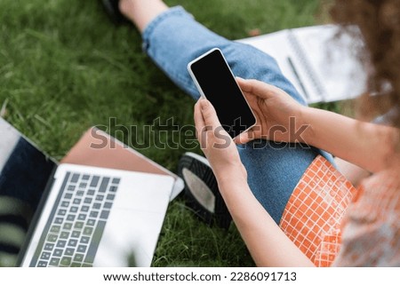cropped view of young woman holding smartphone with blank screen while sitting near laptop and notebook on grass