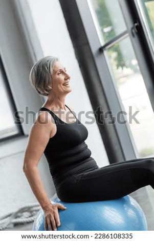 overjoyed senior woman in black leggings and tank top exercising on blue fitness ball