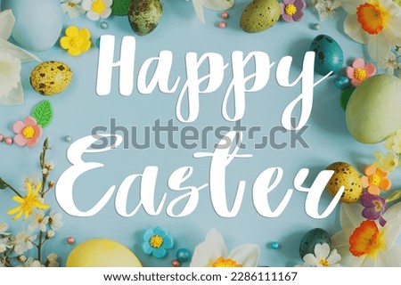 Happy Easter text sign on Easter flat lay with stylish eggs and blooming spring flowers on blue background. Modern Easter greeting card. Handwritten lettering
