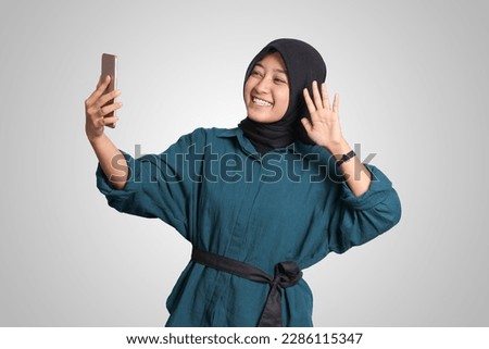 Portrait of excited Asian muslim woman with hijab holding mobile phone while in video calling with her hand gesture saying hi. Advertising concept. Isolated image on white background