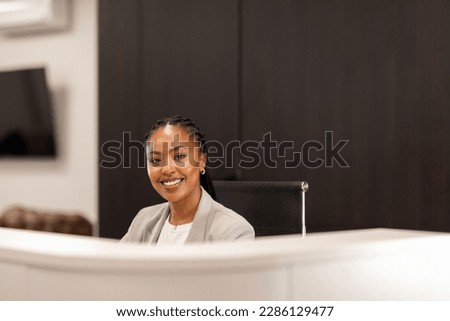 The young African-American female receptionist sits behind the reception desk and smiles at the camera. Royalty-Free Stock Photo #2286129477