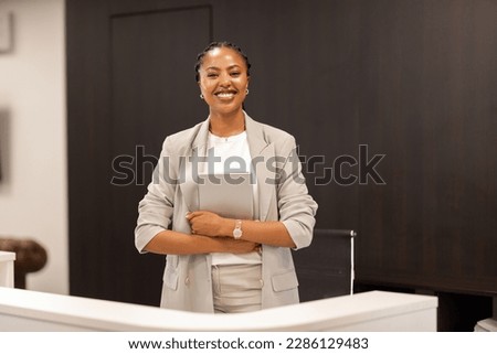 The young African-American female receptionist stands behind the reception desk with a digital tablet in her hands and smiles at the camera. Royalty-Free Stock Photo #2286129483