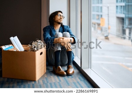 Sad entrepreneur sitting next to box of his belongings and looking through the window after being fired at work. Copy space. Royalty-Free Stock Photo #2286169891