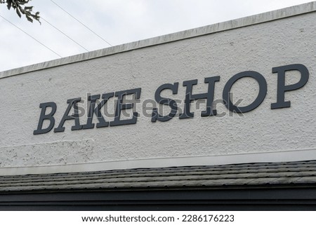 A white building exterior wall of a bake shop with a black colored sign. The text of the bake shop is in capital letters on a baked goods store. The eave of the building has an overhang with a sign.