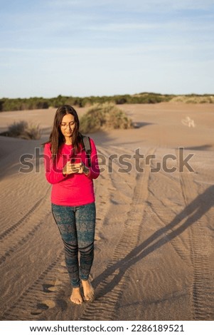 Young bespectacled Latina woman walking face forward using her cell phone on the dunes of a South American beach in Buenos Aires province. Vertical photo