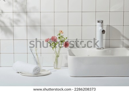 Interior of a modern bathroom with a white tile wall, washstand, towel and toothbrushes. Glass vase with pink flowers that stand out on white background. The concept of bathroom, copy space