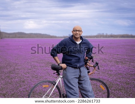 Environmental portrait of smiling  young Indian American man leaning on his bicycles in field of purple wildflowers in spring in Midwest; cloudy blue sky in background