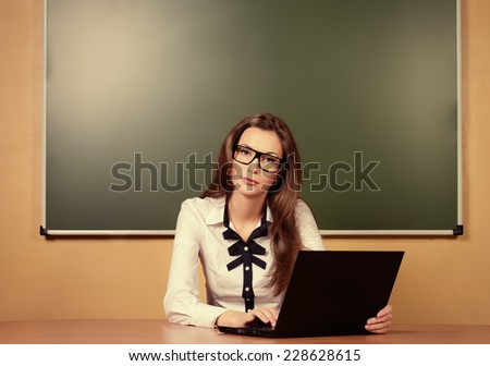 Serious young teacher in spectacles sitting at a desk with a laptop. Education.