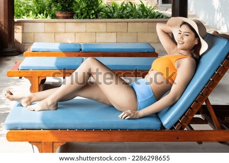 Happy smiling relaxed summer woman wearing bikini swimsuit with hat at swimming pool bench