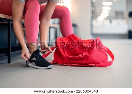 Cropped shot of fit sports woman in sportswear with gym bag wearing pink yoga pants and sneakers sitting on bench, getting ready for exercise session, tying her shoelaces in locker room at gym. Royalty-Free Stock Photo #2286304543