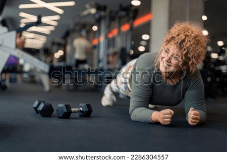 Beautiful happy smiling young woman in comfortable sports clothes feeling motivated determined to work on her body weight to stay active, healthy and in shape practicing plank exercise at gym.