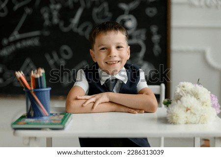 primary school pupil IS SITTING AT HIS DESK AND SMILING AGAINST THE BLACKBOARD BACKGROUND