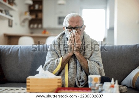 Senior man at home suffering with cold or flu virus. Shot of a senior man blowing his nose with a tissue at home