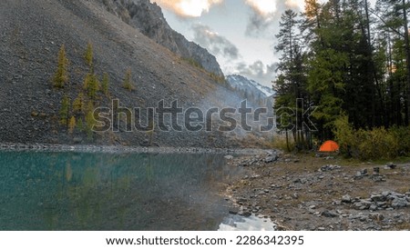 An orange tourist tent stands on the shore of the alpine lake Shavlinskoye near stone cliffs with smoke from a fire in Altai during the day. Royalty-Free Stock Photo #2286342395