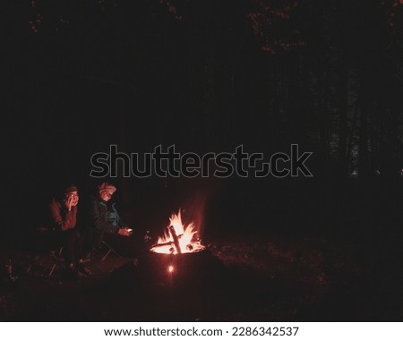 Silhouettes of two friends of a young girl and an elderly woman sitting by a fire in the forest under the trees next to the gazebo in Altai in the dark.
