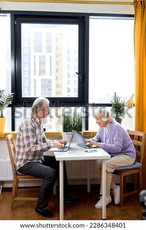 Retired Caucasian People Using Laptop Working Or Studying At Home, Websurfing In Cozy Room. Older Couple Sit Opposite Of Each Other Browsing Internet On Laptop Reading Online News On Weekend