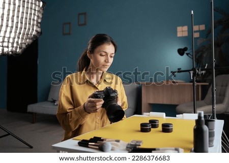 Photographer woman putting cosmetic products on table making composition, making photo of makeup product, holding camera in home studio. Modern blogger. Content creator. Copy space
