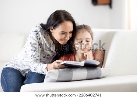 Supportive woman reads a book along with a girl at home on a couch.