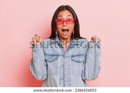 The excited black haired woman stands on light studio background, dressed in denim jacket and pink stylish glasses, raised her hands up and screams with joy,, copy space, high quality photo, enjoyment