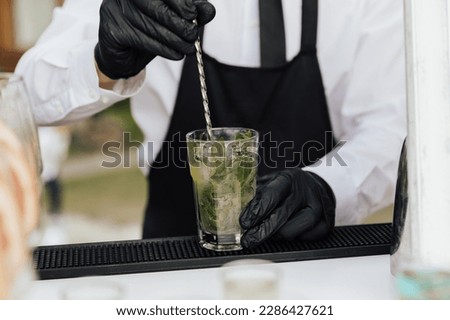 Cropped shot of hands of a professional bartender interfere with a bar spoon the contents of glass with mojito cocktail. Royalty-Free Stock Photo #2286427621