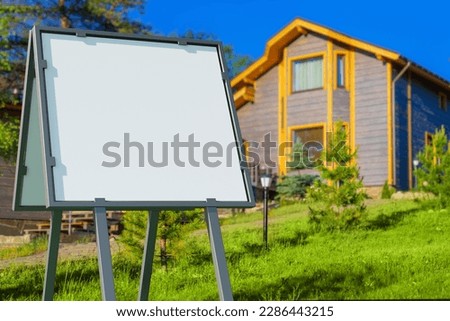 Signboard near house. Two-storey cottage with empty banner. Portable billboard. Signboard for house sale message. Blank advertising sign on lawn of cottage. Template, mockup. Art blurred.