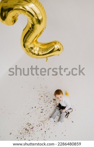 Happy boy having fun celebrating birthday party 2 years old. Confetti. Confetti, falling, flying glitter. Congratulate son on birthday near white wall in studio. Child play with photo camera. Top view
